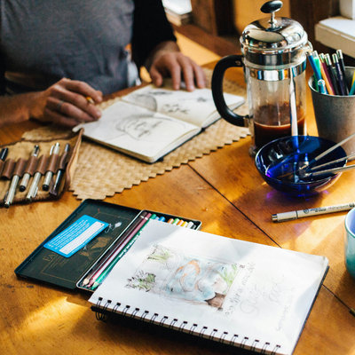 A person seated at a wooden table sketching in an open notebook, surrounded by art supplies including pens, coloured pencils, a roll-up pen case, a bowl with brushes, and a French press of coffee.