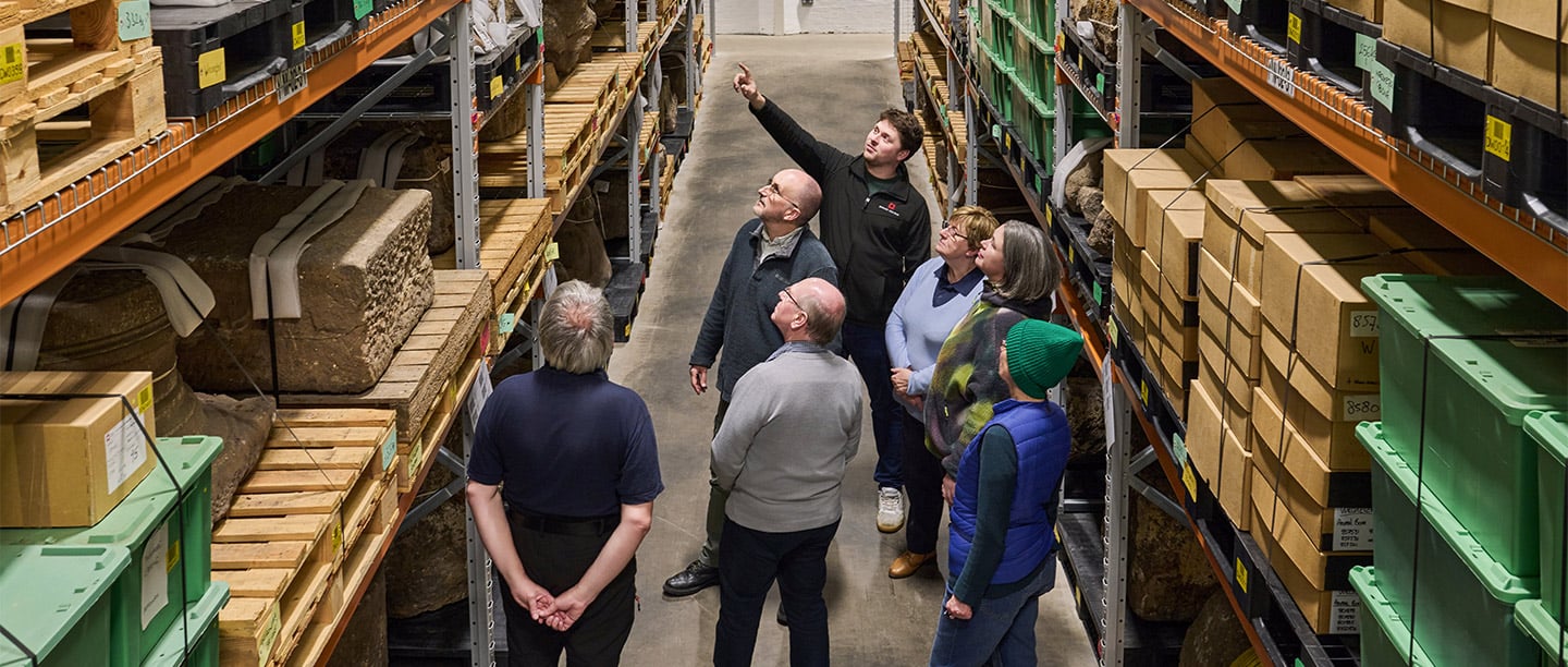 A man stands in front of a group of people in a storeroom pointing at one of the shelves