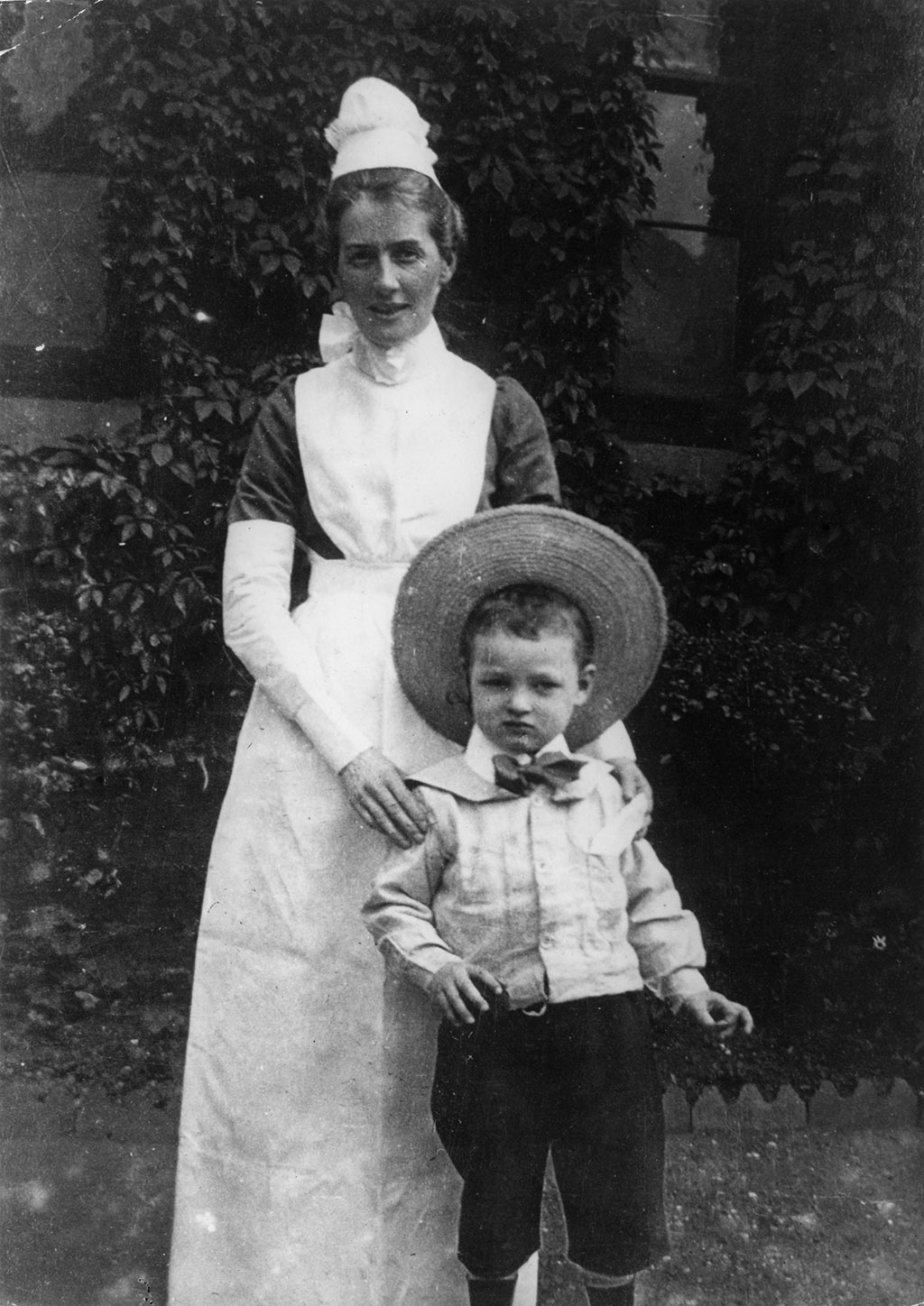 A black and white photograph of Edith Cavell in her nurse's uniform, smiling and standing behind a young child wearing a large straw hat