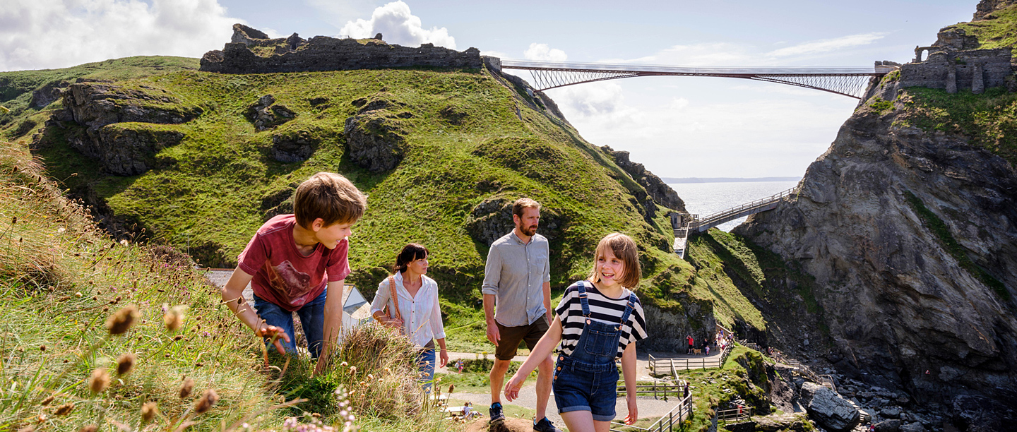 A man and a woman walk behind two children smiling as they walk along the cliffs at Tintagel Castle with the bridge in the background.