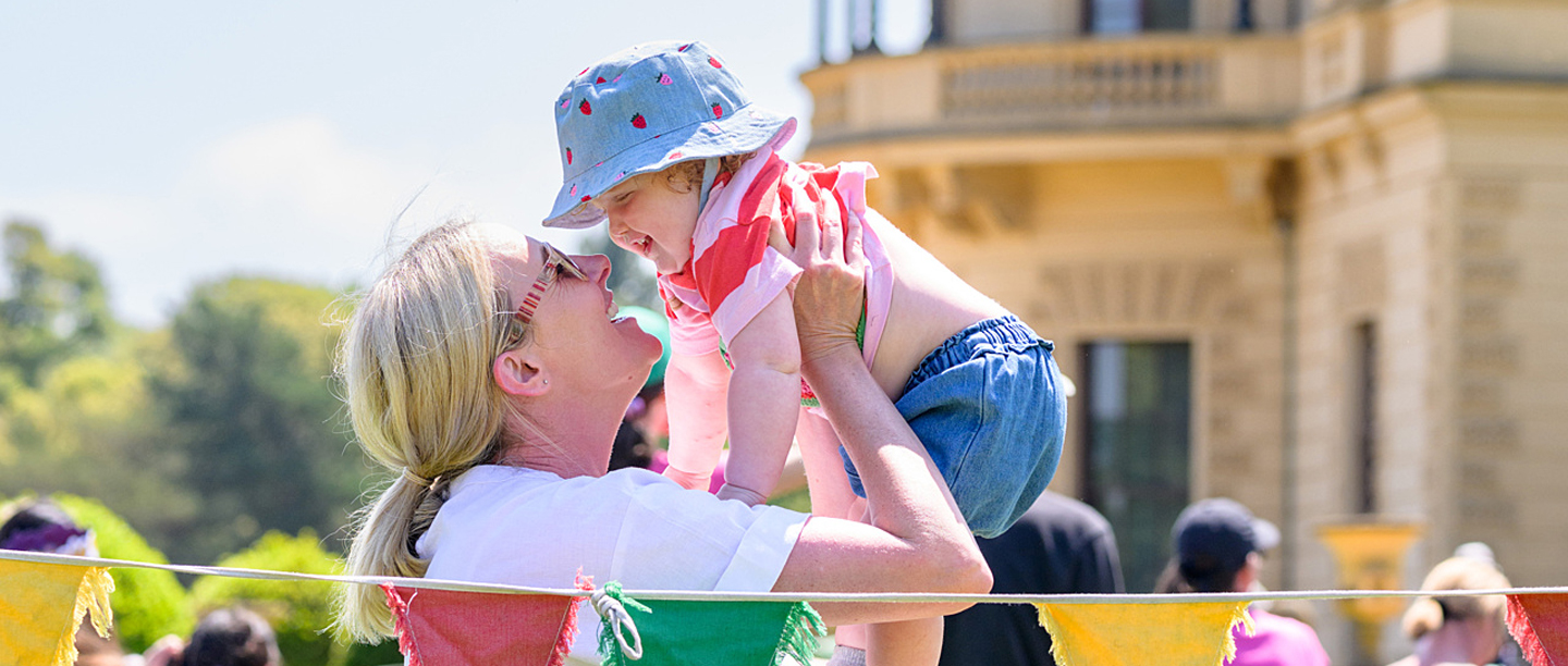 A woman holds a baby up smiling next to a string of colourful bunting with Osborne in the background.