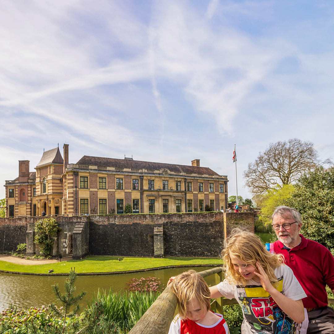 a boy, girl and man walk along the path in the foreground with the moat and Eltham Palace in the background