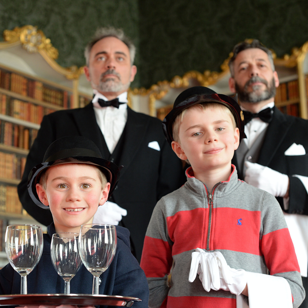 two children in top hats in the foreground, one holds a tray of wine glasses. In the background are two butler's dressed in victorian butler costume.