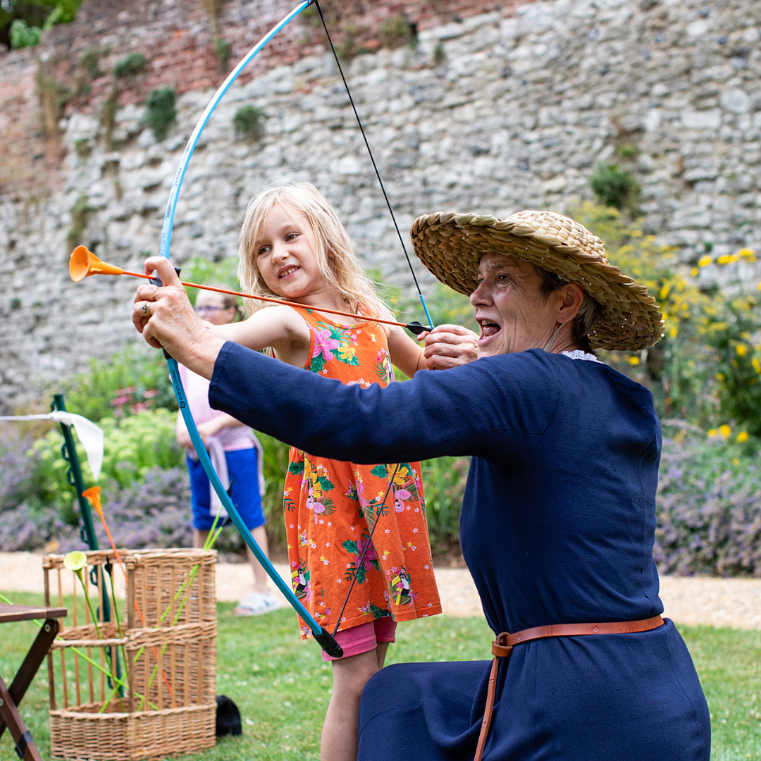 a woman in medieval dress and round hat helps a young girl to use a toy bow and arrow