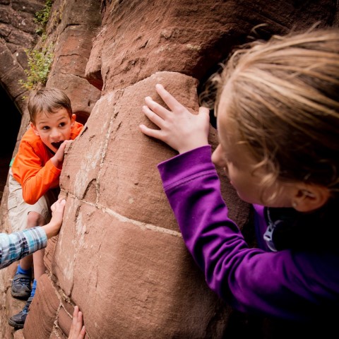 Children climbing and playing within castle walls. A boy in an orange sweatshirt peers behind a stone wall, while a girl in a purple sweatshirt looks at him, gripping the wall. 