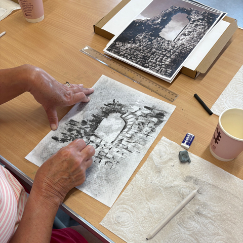 A close-up view of someone drawing a stone ruin with an English Heritage branded coffee cup on the table. 