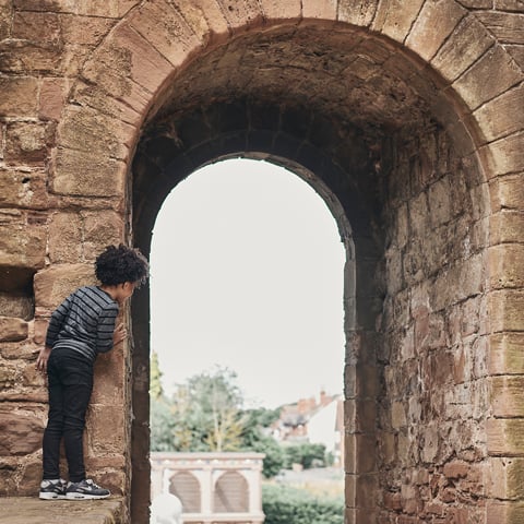 A young boy peers behind a stone archway with view of the gardens at Kenilworth Castle.