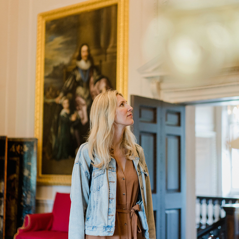 A woman wearing a denim jacket looks up in a room in Marble Hill House. There is a painting of Charles I on the wall behind.