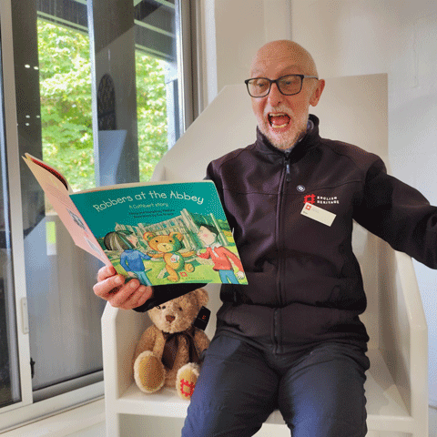 A man sits on a white chair and enthusaistically reads a children's book titled 'Robbers at the Abbey' with his arm raised in motion. He is wearing English Heritage uniform.