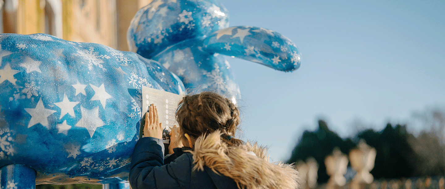 Photo of a young child leaning on a bright-blue statue of Shaun the Sheep to write on a trail map at Wrest Park in Bedfordshire