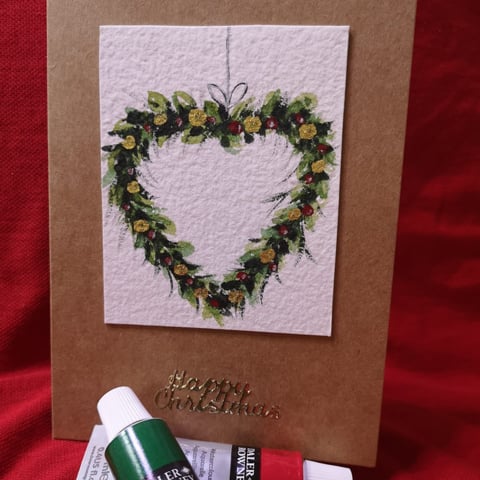 A handmade Christmas card with a small painting of a heart shaped wreath on the front. There are tubes of red and green paint below.
