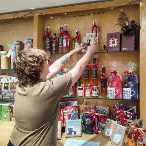 Someone in English Heritage uniform is stacking Christmas themed products on shelves in a gift shop.