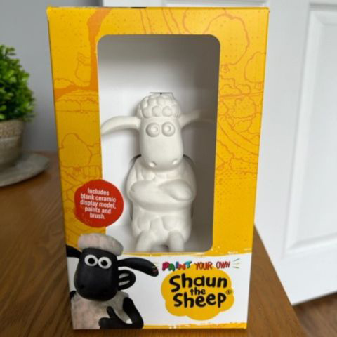 A ceramic Shaun the Sheep model in a yellow and white box on a wooden table.