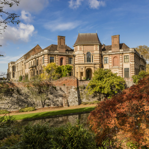 The exterior of Eltham Palace surrounded by trees and a large stone and brick wall and a moat below.