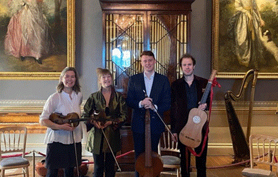 Four people holding string instruments smile for the camera. They are stood in a large room at Kenwood House and are stood infront of two large portraits of women. Four people holding string instruments smile for the camera. They are stood in a large room at Kenwood House and are stood infront of two large portraits of women.