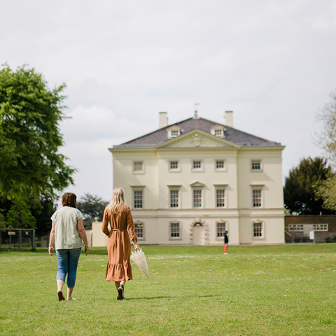 Two women walk towards the front of Marble Hill House in the gardens.
