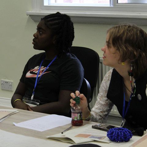 Two young people sit at a table surrounded by papers, pens and books attentively looking toward the left.