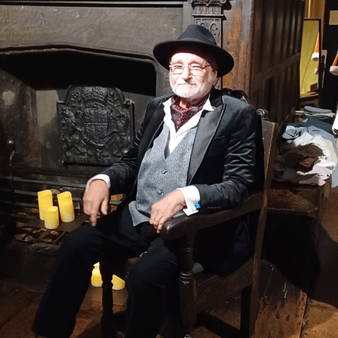 A man dressed in a grey waistcoat, black suit and a black top hat sits on a wooden chair next to lit candles.