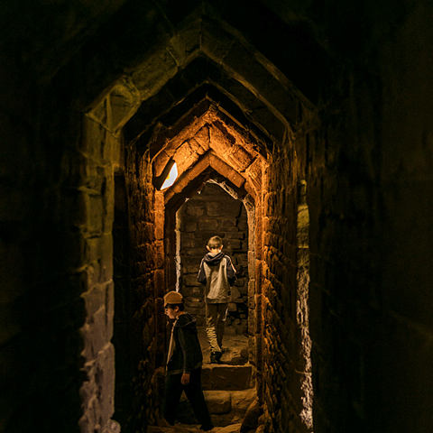 A dimly lit stone castle tunnel with two young children through them. 