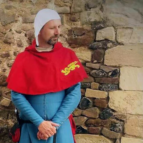 A man dressed in a blue and red medieval costume stands with a sword on his belt.