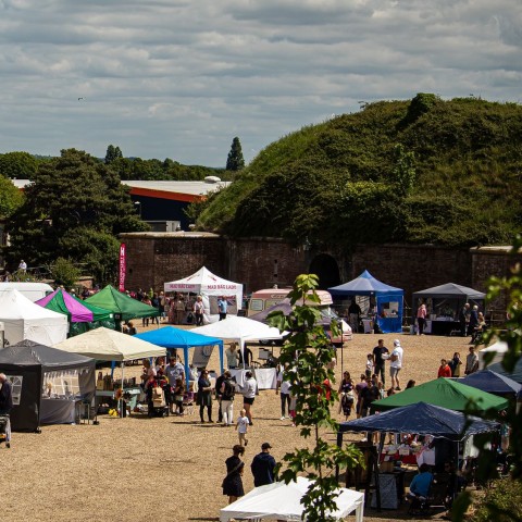 Outdoor craft fair with rows of colourful canopy stalls and visitors in a gravel courtyard beside a red-brick building and trees under a cloudy sky.