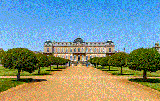 An exterior view of Wrest Park underneath a bright blue sky and rows of trees.  An exterior view of Wrest Park underneath a bright blue sky and rows of trees.