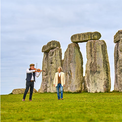 Anton Lesser walking in front of Stonehenge with a musician playing the violin
