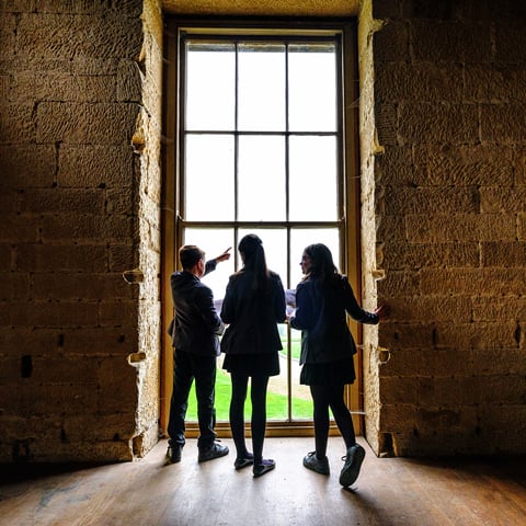 Three children in school uniform look out of a  large window at Belsay Hall.