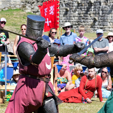 A reenactor dressed in full 13th century armour fights another knight in front of a crowd.