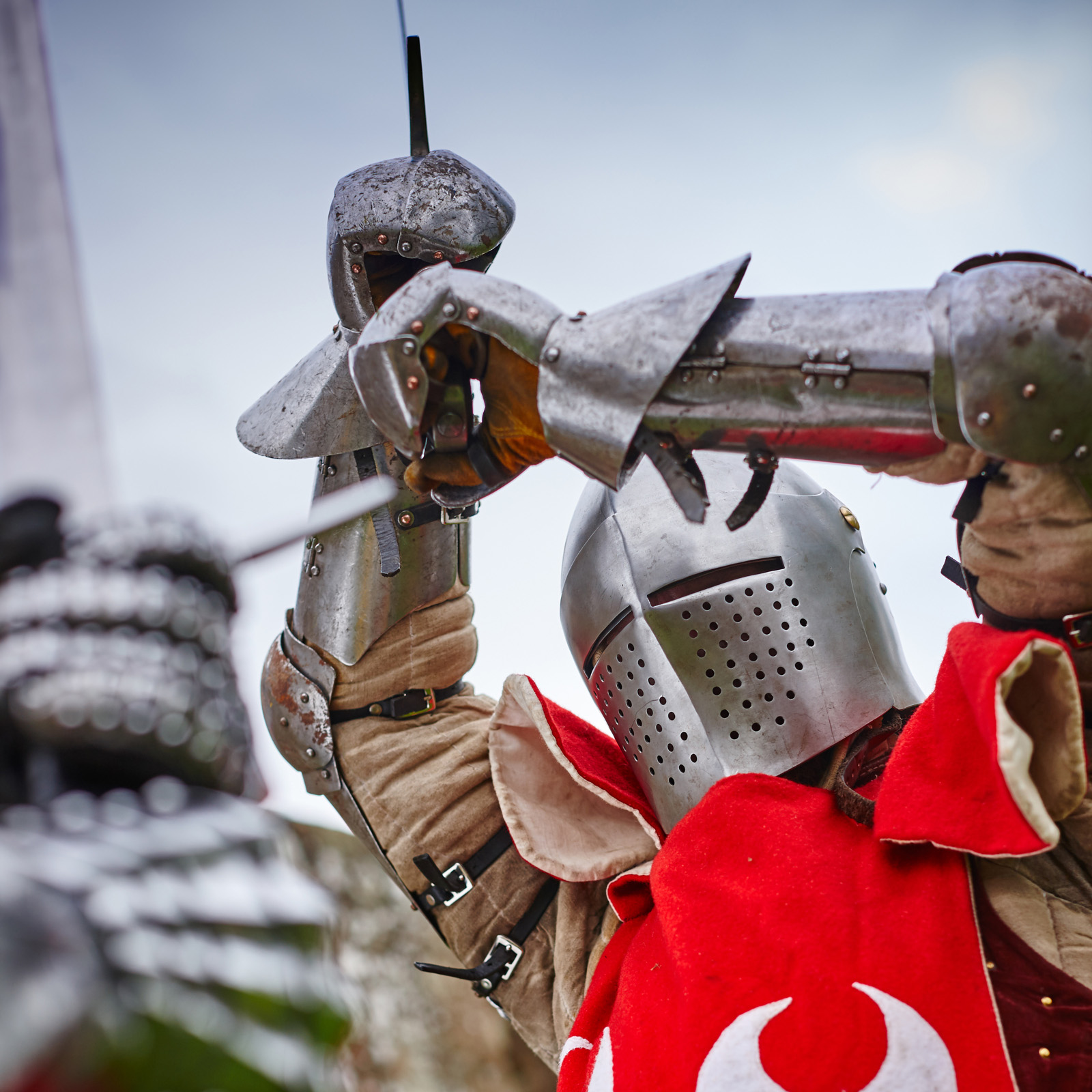 A close-up view of a reenactor dressed a Knight wears full armour and raises a sword.