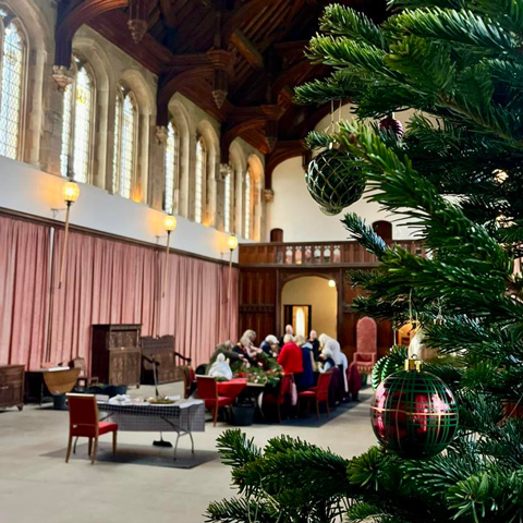 A group of people taking part in a wreath making workshop in the great hall at Eltham Palace with a Christmas tree in the foreground.