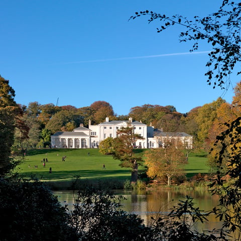 A view of Kenwood House on a sunny day surrounded by greenery and autumn leaves on the trees.