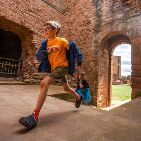 A young boy runs through an archway at Kenilworth Castle with a young girl following behind climbing up stone steps.
