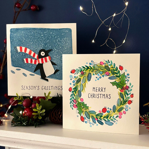 Two handmade watercolour Christmas cards stood on a white wooden fireplace.