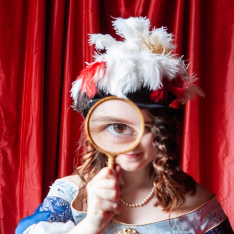 A woman dressed in 17th century costume dress holding a magnifying glass up to her eye.