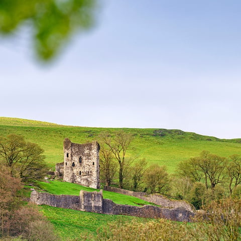 A view from afar of Peveril Castle on a sunny day, surrounded by trees and greenery.