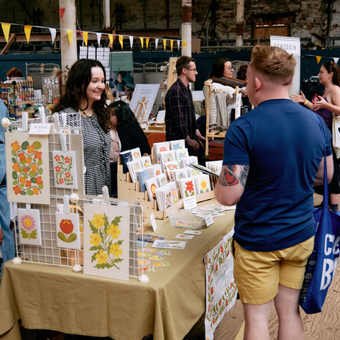 A woman smiles and stands behind a market stall selling colourful prints, engaging with customers.