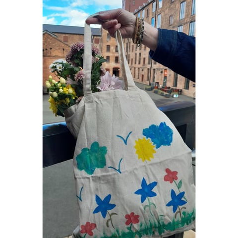 A close-up of someone holding a hand painted fabric tote bag with colourful flowers. Shrewsbury Flaxmill Maltings is in the background.