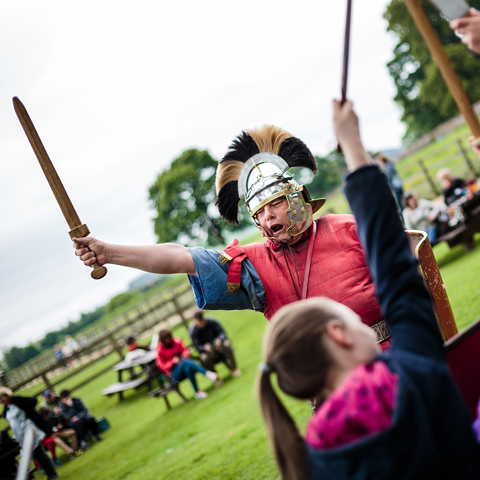 A Roman reeanactor raises a sword infront of a young child copying the action.