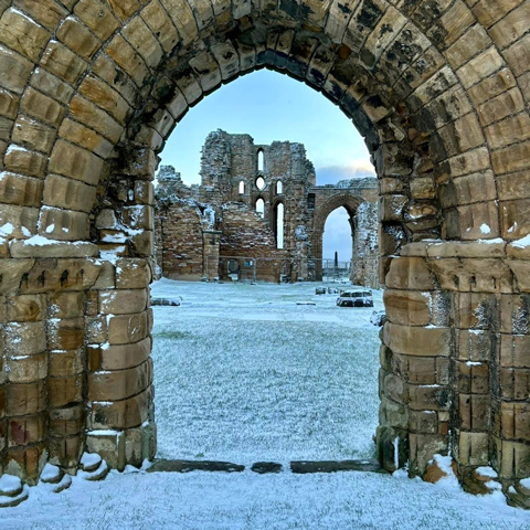 A view of Tynemouth Priory ruins covered in snow through a stone archway.