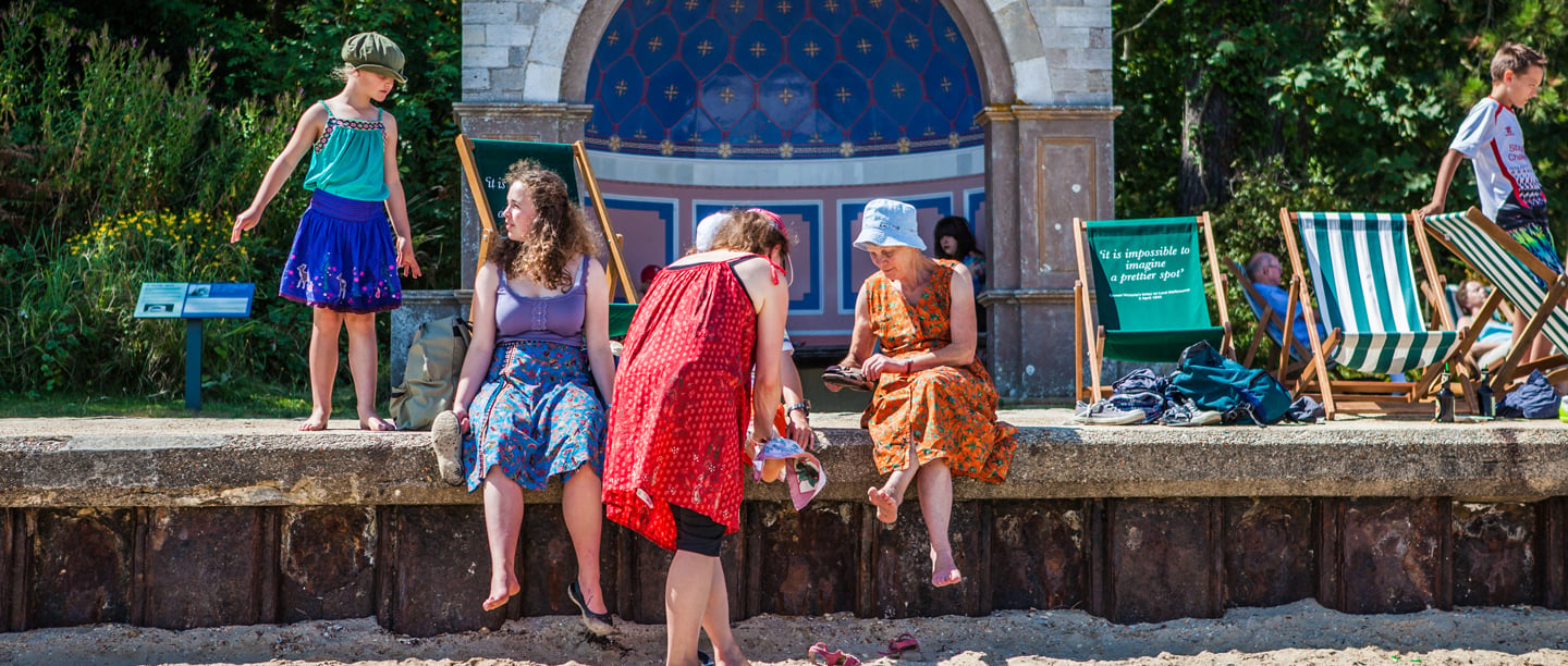 A group of adults and children sitting and standing on the beach at Osborne dressed in summer outfits.