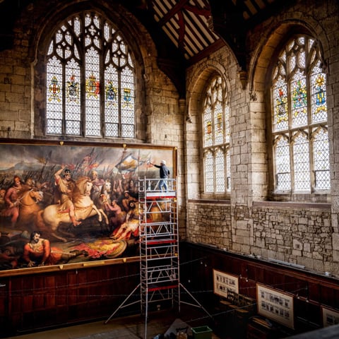 A conservator stands on scaffolding of a large painting in the Abbot's Hall at Battle Abbey.