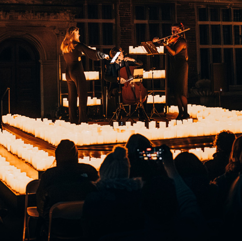 A string and woodwind band perform on a stage at candlelight in front of a seated audience.