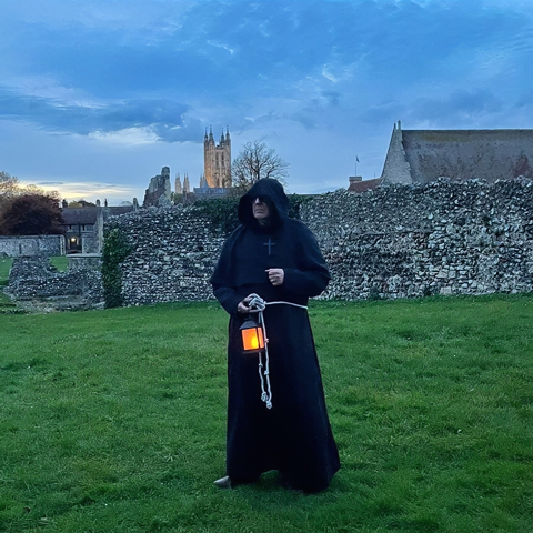 A man dressed in a black monk's cloak stands with a lanterns on the grounds of St Augustine's Abbey 