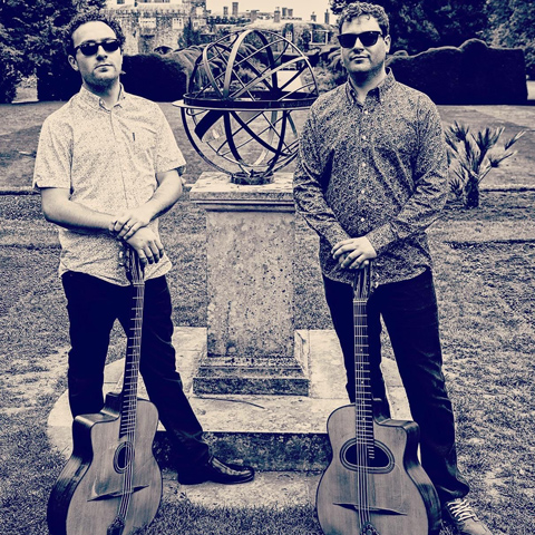 A sepia image of the Barrett Brothers standing by acoustic guitars in gardens in front of a globe like sculpture.