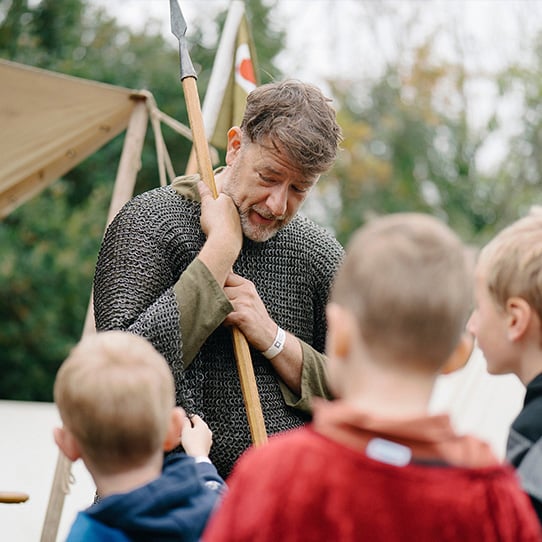 A reenactor Knight wearing chainmail stands in front of two young children. 