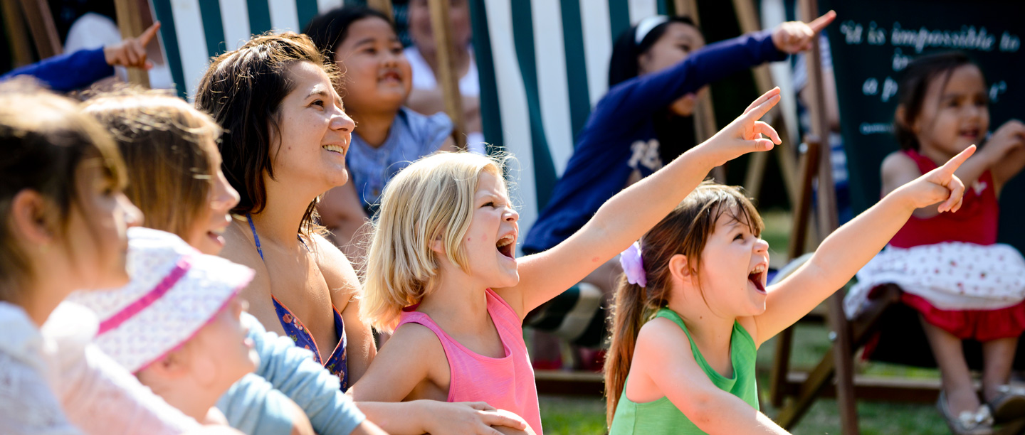 A group of children sat by adults point and shout at a puppet show at Osborne on a sunny day.