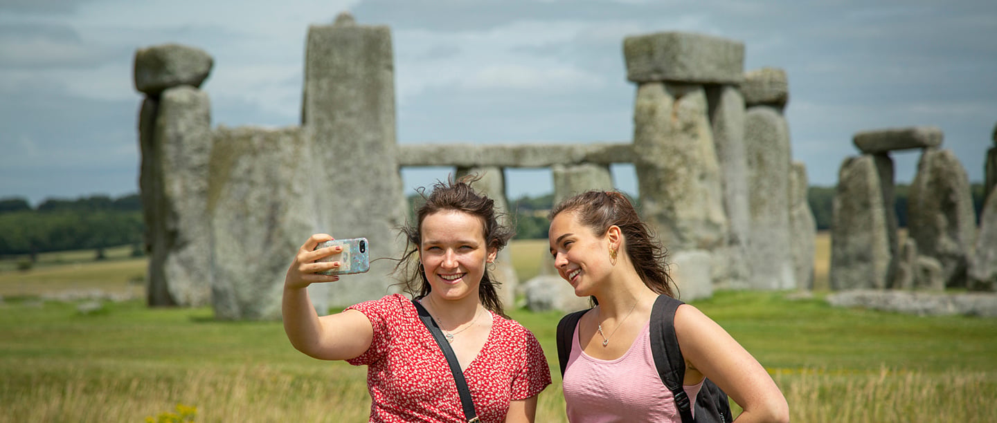 Two young adults pose for a selfie in front of Stonehenge on a cloudy day.