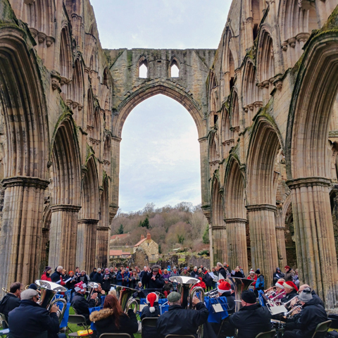 A brass band play to an audience in the middle of the ruins at Rievaulx Abbey.